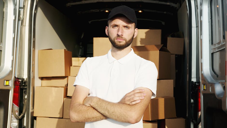 A young man with a serious expression, wearing a white polo shirt and black cap, stands with his arms crossed in front of an open van loaded with cardboard boxes of various sizes, some wrapped in plastic or fabric padding. The boxes are stacked inside the vehicle, which appears to be parked outside a building, with visible pavement and the building's doorway in the background. The scene depicts the loading process involved in home relocation or furniture transport, with the man representing a professional mover or removal expert from Movers Kennington. Bright natural lighting illuminates the interior and exterior environment, emphasizing the packing and loading stage of the moving service.