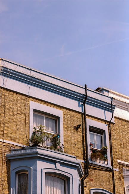 A multi-storey residential building with a brick facade and white window frames, featuring two windows on the upper floor with flower boxes and window curtains. Outside, there is a black downspout pipe running vertically along the wall, and the building has a small balcony with a white railing and potted plants. The sky is clear and blue, indicating daytime. Although the image does not show actual moving activities or furniture, it reflects the exterior environment of a typical home suitable for house removals and relocation services, as provided by Movers Kennington. This scene highlights the urban residential setting where furniture transport and packing preparations could take place during a home relocation process, with natural daylight emphasizing the structural details of the property.