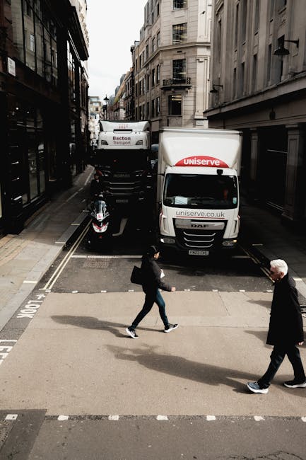 A street scene showing the loading area of a house removal process, with a white moving truck marked with 'uniserve' and 'uniserve.co.uk' parked across a narrow alleyway. The truck is partially blocking the pavement while two large vehicles are parked behind it, one with the label 'Becton Transport.' In the foreground, a pedestrian with a black backpack and dark clothing is crossing the road, while another person with white hair, dressed in dark clothing, is walking in the opposite direction. The surrounding environment includes multi-storey buildings with narrow facades, typical of urban residential areas, with some windows visible. The setting appears to be during the daytime with natural light illuminating the scene, capturing a moment within a home relocation and furniture transport context involving loading or unloading activities, as part of a professional house removal service in Kennington, operated by Movers Kennington.