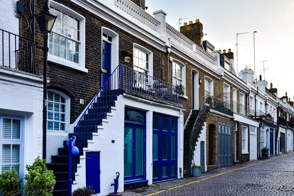 A row of terraced houses with brick facades and white trims on a cobblestone street in Kennington. Each property features a ground-floor entrance with a large glass door framed in blue, and small front gardens or planters at the base of the stairs. The houses have external staircases with blue railings leading to second-floor entrances, some with potted plants on the balconies. Visible windows have white casings, with some open or partially covered with blinds. Moving equipment, such as trolleys and packing materials, are not present, but the setting suggests preparation for a home relocation involving furniture transport or packing. The street appears quiet with soft natural lighting, and the overall scene reflects typical residential logistics involved in home removals in Kennington, with Movers Kennington potentially preparing for or documenting a house move involving careful loading and staging of belongings outside the property.
