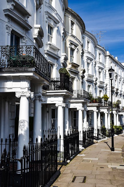 A row of white Victorian terraced houses in Kennington with ornate architectural details, including decorative cornices and black wrought iron balconies on the upper floors. The buildings have large windows and some feature potted plants on the balconies. The pavement in front of the houses is clean and paved with light-colored stones, separated by black metal railings that run alongside the sidewalk. The sky is clear with a few clouds, and street lamps are visible along the walkway. Inside one of the houses, movers from Movers Kennington are preparing for a home relocation by packing furniture and household items into cardboard boxes wrapped with plastic film, and using furniture blankets and straps for protection during the loading process. They are transferring items from inside the property to a nearby van, which is partially visible on the street, ready for transport. The scene emphasizes the logistics of household moving services associated with house removals in the area, highlighting the careful handling of furniture and packing materials involved in a typical house removal operation.