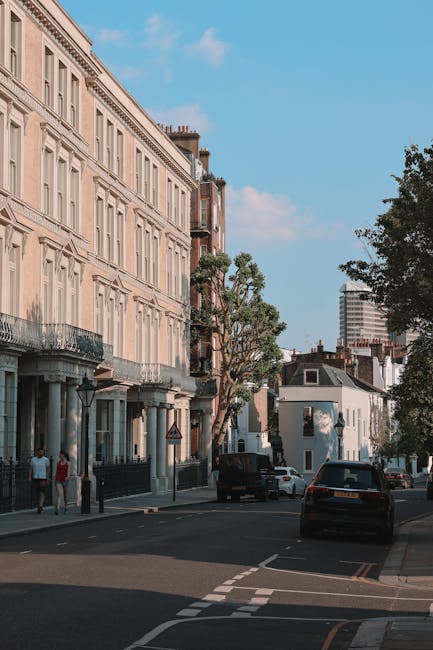 A row of terraced houses with brick facades and white trims on a cobblestone street in Kennington. Each property features a ground-floor entrance with a large glass door framed in blue, and small front gardens or planters at the base of the stairs. The houses have external staircases with blue railings leading to second-floor entrances, some with potted plants on the balconies. Visible windows have white casings, with some open or partially covered with blinds. Moving equipment, such as trolleys and packing materials, are not present, but the setting suggests preparation for a home relocation involving furniture transport or packing. The street appears quiet with soft natural lighting, and the overall scene reflects typical residential logistics involved in home removals in Kennington, with Movers Kennington potentially preparing for or documenting a house move involving careful loading and staging of belongings outside the property.