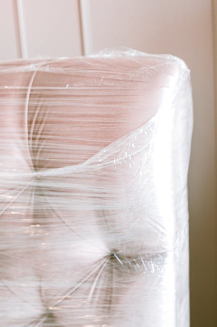 A large, wrapped piece of furniture, possibly a wardrobe or large box, secured with transparent plastic wrap for protection during a home relocation. The furniture is placed inside a room, near a door