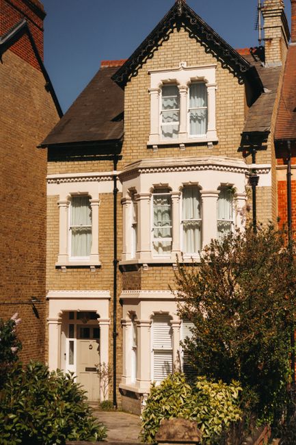 A close-up view of a two-story brick house facade showing two windows on the upper floor, each with white uPVC frames and partially closed beige blinds. Below, there is a small bay window with three s