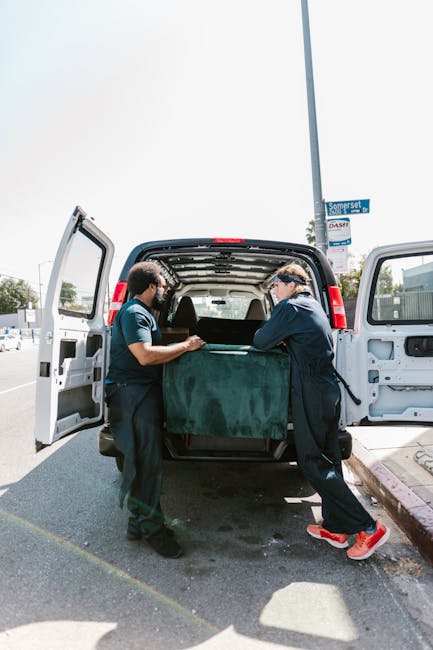 Two professional movers from Movers Kennington are engaged in a home relocation task, loading a large, dark green upholstered furniture piece into the rear of a white moving van parked on a paved street. The interior of the van is visible with the open rear doors, revealing a spacious cargo area prepared for furniture transport. One mover, wearing a blue T-shirt and dark pants, is grasping the furniture from one side, while the other, dressed in a dark jacket and red sports shoes, is assisting from the opposite side. The scene is set outdoors during daylight, with a street sign and urban surroundings, including parked cars and commercial buildings, in the background. The vehicle is positioned close to the curb, with the loading process supported by equipment such as moving blankets and possibly lifting straps, although these are not clearly visible. This image illustrates the logistics involved in furniture packing and loading during a house removal, emphasizing careful handling and professional service to ensure safe transport of belongings, consistent with the services offered by Movers Kennington for residential moves in the SE11 area.