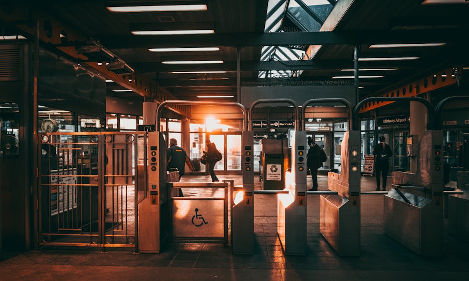 The image shows the interior of a train station entrance during sunset, with multiple entry gates, including some designated for disabled access with a wheelchair symbol. Several passengers are walking through the gates, carrying luggage and backpacks. The station features a modern design with glass doors allowing natural light to illuminate the space. Inside, there are packaging materials, such as cardboard boxes and protective coverings, placed near the entrance areas, indicating preparations for home relocation or moving activities. The lighting creates a warm glow, casting long shadows across the tiled floor, and the station's signage is visible in the background. This setting exemplifies a typical environment for busy relocation logistics, where carefully managed loading, baggage handling, and transportation are essential, consistent with services offered by movers like Movers Kennington for house removals and furniture transport in the area. The image reflects the process of packing, loading, and moving furniture in a well-organized station environment.