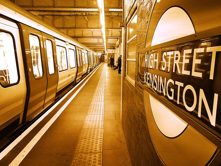 Inside a London underground station, a group of commuters and travelers are walking along the platform between two trains. The platform has tactile paving and safety lines near the edge, with yellow and black markings. Some individuals carry backpacks, suitcases, and shopping bags, and a few are wearing face masks. The station features a curved ceiling with integrated lighting and a staircase leading to the surface or another level. The scene captures the typical flow of people during home relocation or moving processes, reflecting meticulous packing, furniture transport, and logistical planning associated with house removals. This setting is relevant for illustrating access tips for flat removals in Kennington, as provided by Movers Kennington, as they assist with loading and unloading in urban environments.