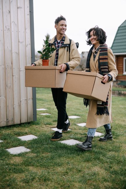 Two movers from Movers Kennington are outdoors, walking across a grassy garden area carrying large cardboard boxes, likely for home relocation purposes. The male mover on the left is wearing a beige coat, checkered shirt, and dark trousers, smiling as he holds a box with both hands. The female mover on the right is dressed in a long beige coat, patterned scarf, and dark boots, also smiling while carrying a similar box. They are approaching a wooden door frame on the side of a house, with a wooden fence and another house with a green roof visible in the background. The boxes are made of sturdy cardboard, and the scene suggests an organized packing and loading process as part of a professional removal service. Natural daylight illuminates the scene, with the movers preparing for furniture transport or packing and moving activities related to house removals on the Myatt Garden Estate.
