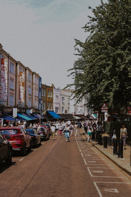 A street scene showing a row of multi-storey buildings with shop fronts and awnings along a paved road in a busy urban area. Parked cars line the left side of the street, while pedestrians walk along the pavement and across the road. Large green trees extend over the sidewalk, providing shade, and a white triangular road sign is visible near the curb. The overall scene suggests a lively shopping district or town centre during daytime. The image, associated with moverskennington.co.uk, relates to house removals and home relocation services, illustrating the logistics involved in moving furniture and boxes through urban environments, with a focus on loading and transportation processes.