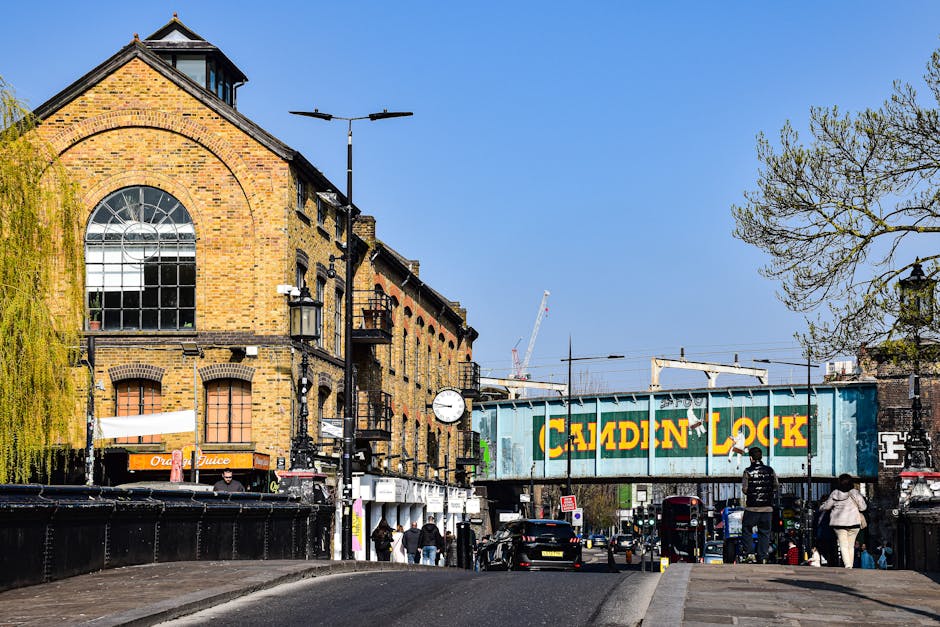 A city street scene in Kennington SE11 showing a brick building with large arched windows and a shop sign for 'Orange Juice' on the ground floor. In the foreground, there are pedestrians walking along the pavement, some carrying bags, and a partly visible black van or container. A prominent railway bridge with a blue and yellow 'CADDIN LOCK' sign spans the street, and behind it, there are parked cars and a moving crane in the background. The street is lined with lampposts, trees, and other urban infrastructure, with a clear blue sky overhead. The scene captures a typical day of urban activity, with vehicles, walking pedestrians, and ongoing city transport and logistical operations, supporting the context of house removals and home relocation activities such as furniture transport and packing within the area served by Movers Kennington.